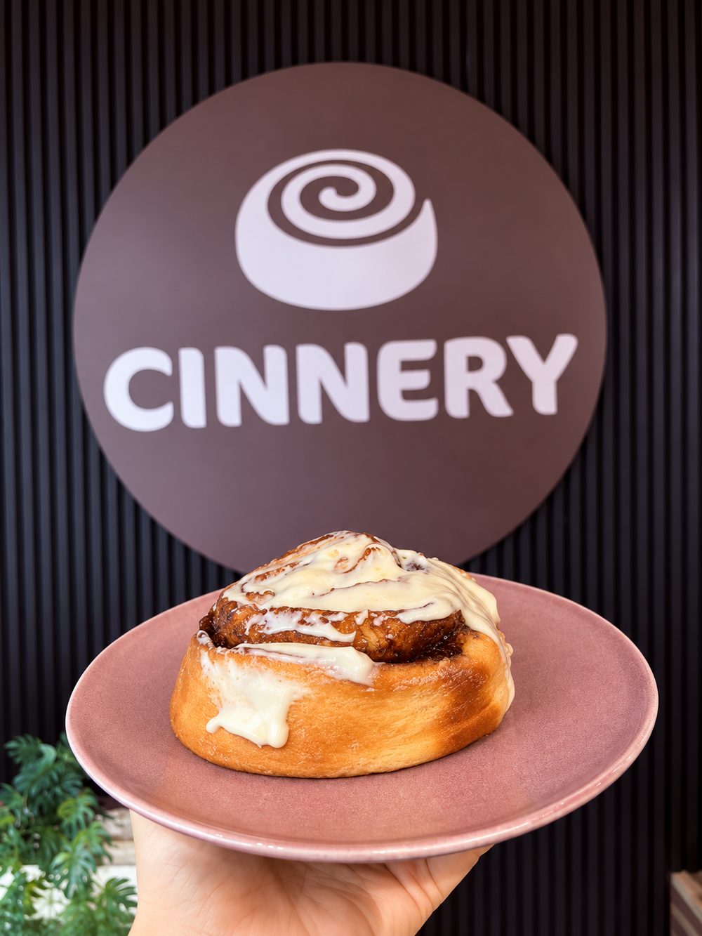 Cinnamon roll on a pink plate in front of the Cinnery sign